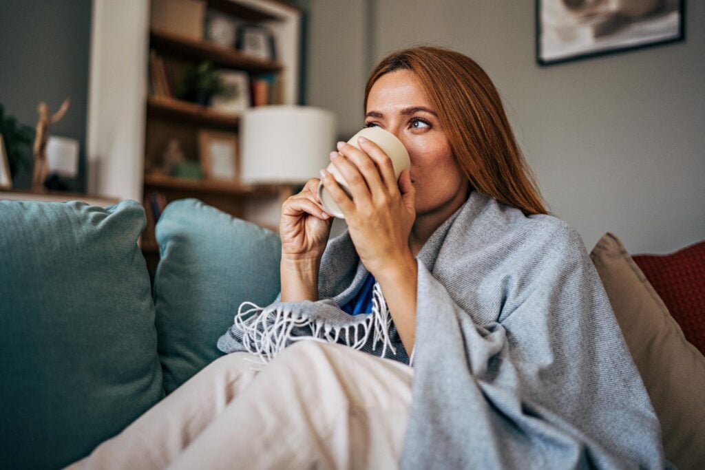 Kvinna sitter under en filt i soffan och dricker kaffe